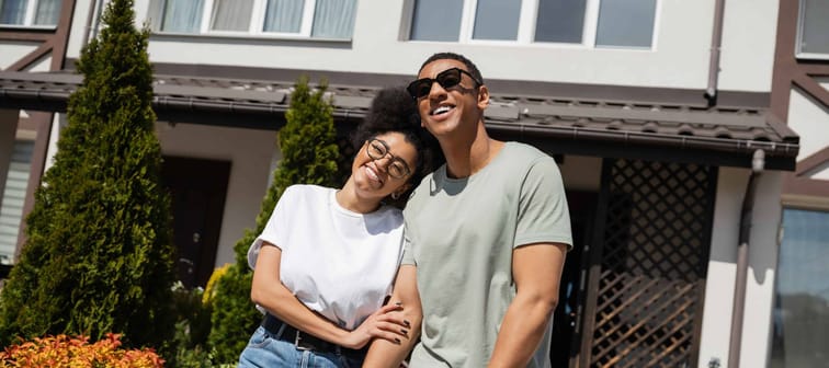 smiling African-American woman in eyeglasses hugging boyfriend near new house at background